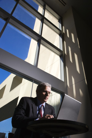 Low angle view of a businessman with a laptop computer in front of large windows. Vertical shot.